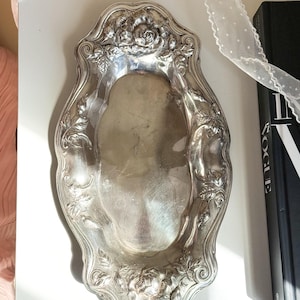 Puede incluir: Bandeja plateada ornamentada con un diseño floral y de hojas en los bordes. La bandeja tiene forma ovalada alargada con un borde ligeramente elevado. Los detalles florales incluyen rosas y otros elementos decorativos.