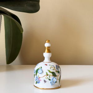 Puede incluir: Una campana de porcelana blanca con asa y borde dorados. La campana est&aacute; decorada con dise&ntilde;os florales azules y vides verdes, junto con peque&ntilde;os detalles de insectos rojos y negros.