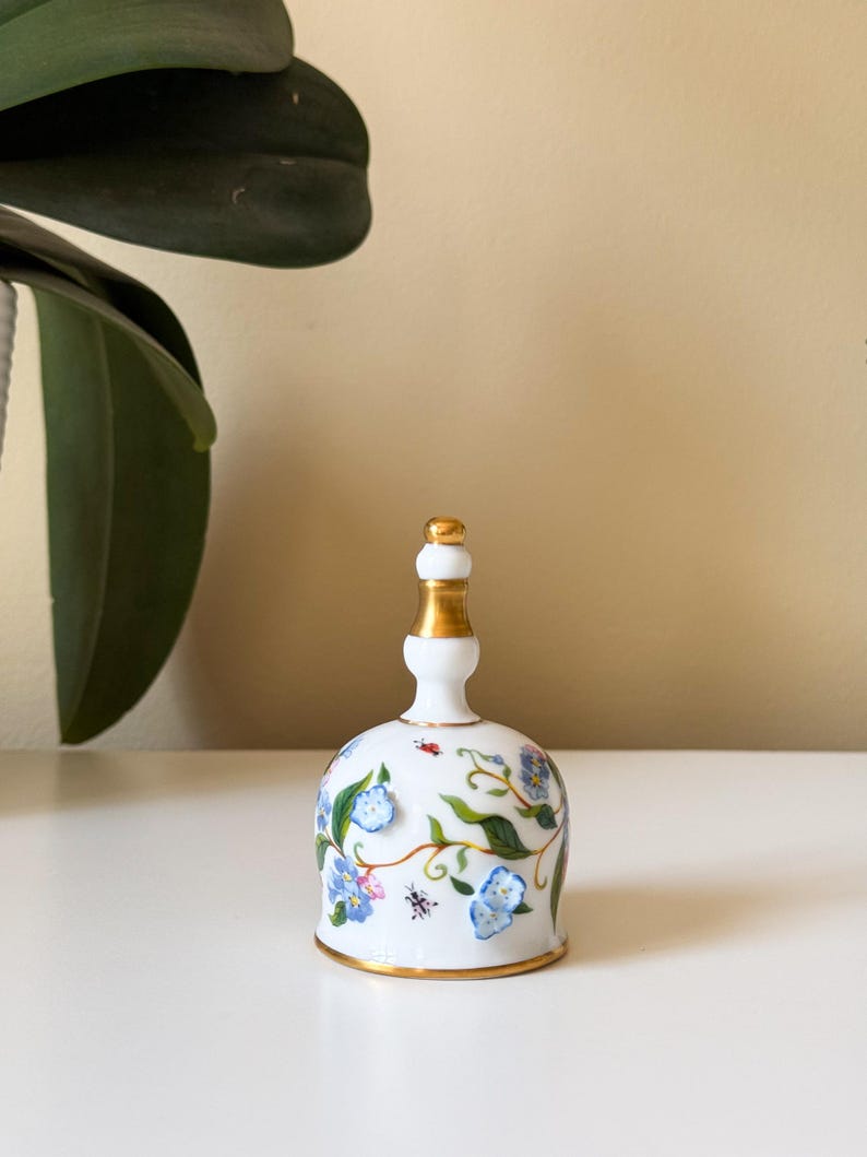 Puede incluir: Una campana de porcelana blanca con asa y borde dorados. La campana est&aacute; decorada con dise&ntilde;os florales azules y vides verdes, junto con peque&ntilde;os detalles de insectos rojos y negros.
