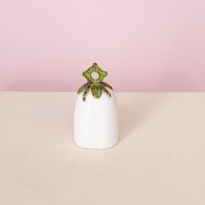 Puede incluir: Una campana de cer&aacute;mica blanca con un asa en forma de hoja verde y dorada. La campana tiene un acabado liso y brillante y una base ligeramente acampanada. El asa est&aacute; detallada con hojas verdes y detalles dorados.