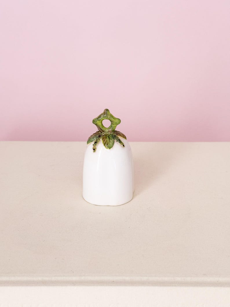Puede incluir: Una campana de cer&aacute;mica blanca con un asa en forma de hoja verde y dorada. La campana tiene un acabado liso y brillante y una base ligeramente acampanada. El asa est&aacute; detallada con hojas verdes y detalles dorados.