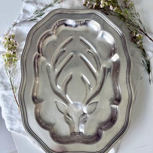 May include: Silver-toned metal serving platter with a raised deer head design in the center. The platter has a scalloped edge and is set on a white cloth with sprigs of small flowers.