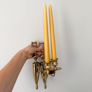 May include: A vintage brass wall sconce with two yellow taper candles. The sconce has a decorative design with a teardrop shape and a circular candle holder. The candles are approximately 10 inches tall.