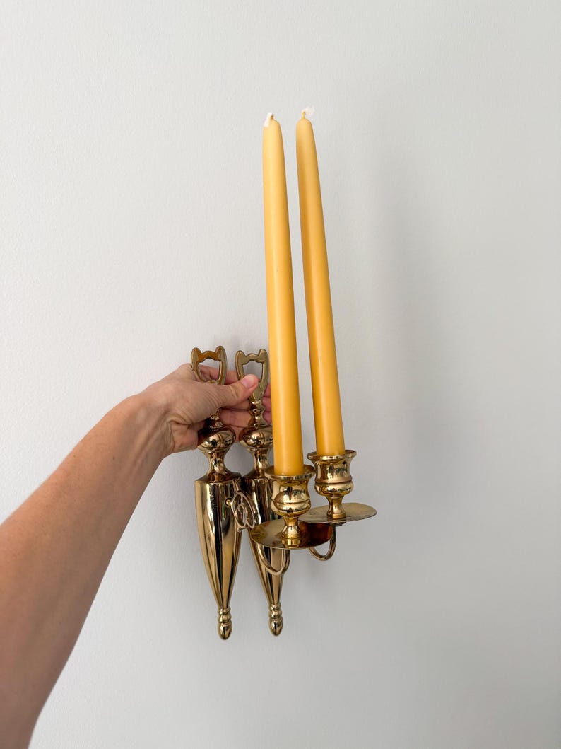 May include: A vintage brass wall sconce with two yellow taper candles. The sconce has a decorative design with a teardrop shape and a circular candle holder. The candles are approximately 10 inches tall.