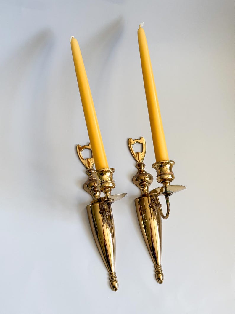 May include: Two vintage brass wall sconces with yellow candles. The sconces have a decorative design with a conical base and a curved arm to hold the candle. The candles are tall and slender.
