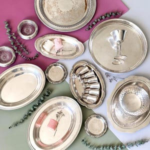 May include: An assortment of vintage silver serving trays and dishes, including oval platters, round trays, and small coasters. Some trays have ornate detailing, and a few contain decorative items like flowers and ribbon. The items are arranged on a pink and green surface.