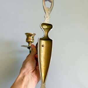 May include: A vintage brass wall sconce with a candle holder and a decorative backplate. The sconce has a gold finish with a detailed design. The candle holder is designed to hold a single candle. The sconce is held by a hand.