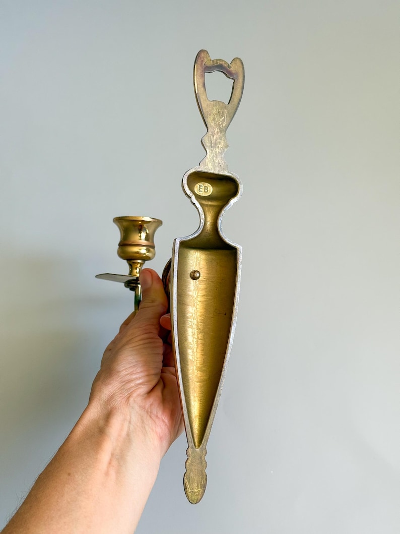May include: A vintage brass wall sconce with a candle holder and a decorative backplate. The sconce has a gold finish with a detailed design. The candle holder is designed to hold a single candle. The sconce is held by a hand.