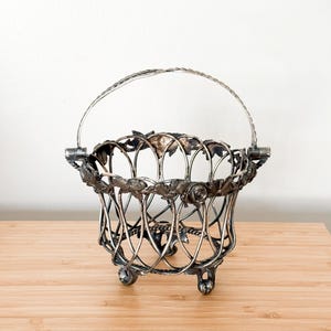 May include: A silver-toned metal basket featuring a lattice-style wire body, a hinged handle, and three small feet. The upper rim is decorated with a relief pattern of leaves and roses, and the metal has a darkened, aged patina finish.