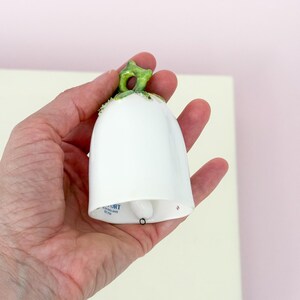 Puede incluir: Una campana de cer&aacute;mica blanca con un tallo verde y un peque&ntilde;o badajo negro. La campana se sostiene en una mano sobre un fondo rosa claro y blanco roto. La campana tiene la palabra "PORT" impresa.