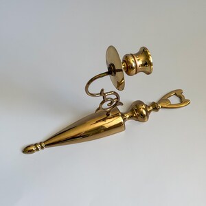 May include: A vintage brass candlestick holder and bottle opener set. The candlestick holder features a round base, curved arm, and a cup for the candle. The bottle opener has a long, pointed body with decorative details and a bottle opener end.