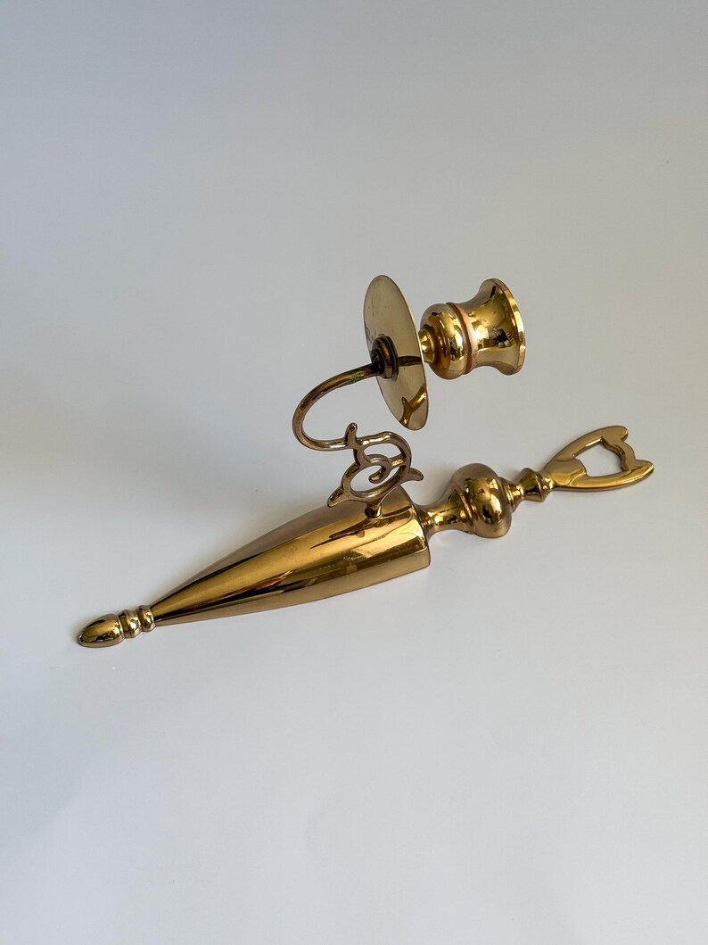 May include: A vintage brass candlestick holder and bottle opener set. The candlestick holder features a round base, curved arm, and a cup for the candle. The bottle opener has a long, pointed body with decorative details and a bottle opener end.