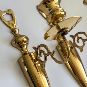 May include: A pair of vintage brass wall sconces. The sconces have a polished gold finish and feature a candle holder and decorative scrollwork. The sconces are designed to hold candles and add a touch of elegance to any room.