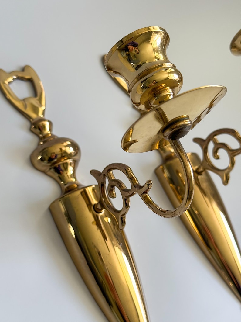 May include: A pair of vintage brass wall sconces. The sconces have a polished gold finish and feature a candle holder and decorative scrollwork. The sconces are designed to hold candles and add a touch of elegance to any room.