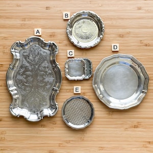 May include: An assortment of antique silver serving trays and dishes. The collection includes a large rectangular tray with ornate detailing, a small round dish with a floral design, and a square dish. The items are displayed on a wooden surface.