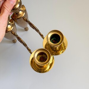 May include: Two vintage brass candle holders with ornate handles. The candle holders have a classic design with a round base and a cup for holding candles. The brass has a warm, golden tone, and the handles have a twisted design.