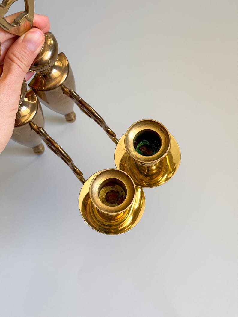 May include: Two vintage brass candle holders with ornate handles. The candle holders have a classic design with a round base and a cup for holding candles. The brass has a warm, golden tone, and the handles have a twisted design.