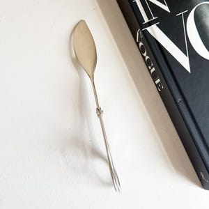 May include: A silver-colored serving utensil with a leaf-shaped head and a long, thin handle. The handle has a loop at the end. The utensil is next to a black book with the words "IN VOGUE" in white.