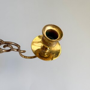 May include: A vintage brass candlestick holder with a circular base and a cup-shaped top. The candlestick has a curved arm with decorative cutouts. The brass has a warm, golden hue, and the interior of the cup is dark.