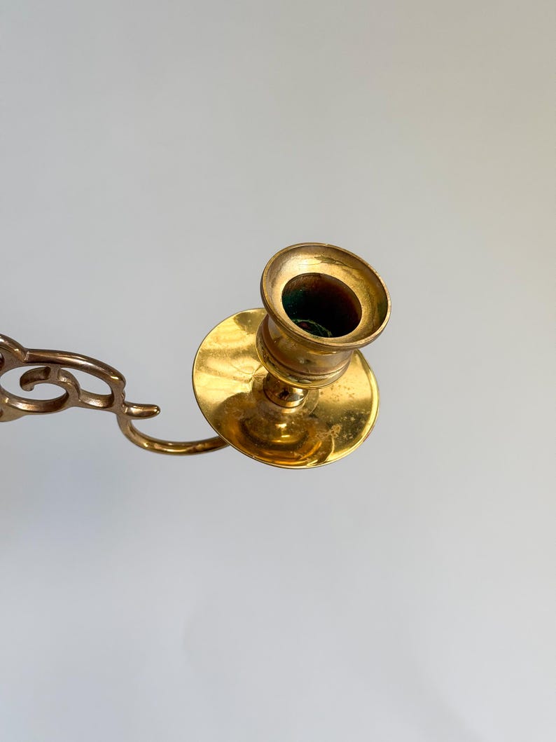 May include: A vintage brass candlestick holder with a circular base and a cup-shaped top. The candlestick has a curved arm with decorative cutouts. The brass has a warm, golden hue, and the interior of the cup is dark.