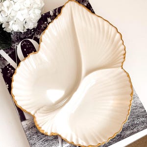 Può includere: Un piatto da portata a forma di foglia color crema con bordo dorato. Il piatto ha un design diviso e una superficie strutturata e a coste. Il piatto è appoggiato su una rivista con una fotografia in bianco e nero.