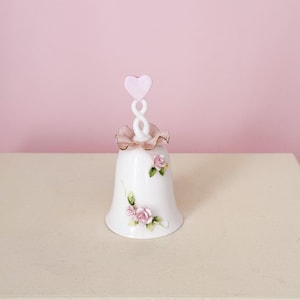 Puede incluir: Campana de porcelana blanca con detalles florales de rosas rosas. La campana tiene un asa en forma de coraz&oacute;n rosa y un ribete con volantes. Un art&iacute;culo decorativo, probablemente para exhibici&oacute;n o colecci&oacute;n.