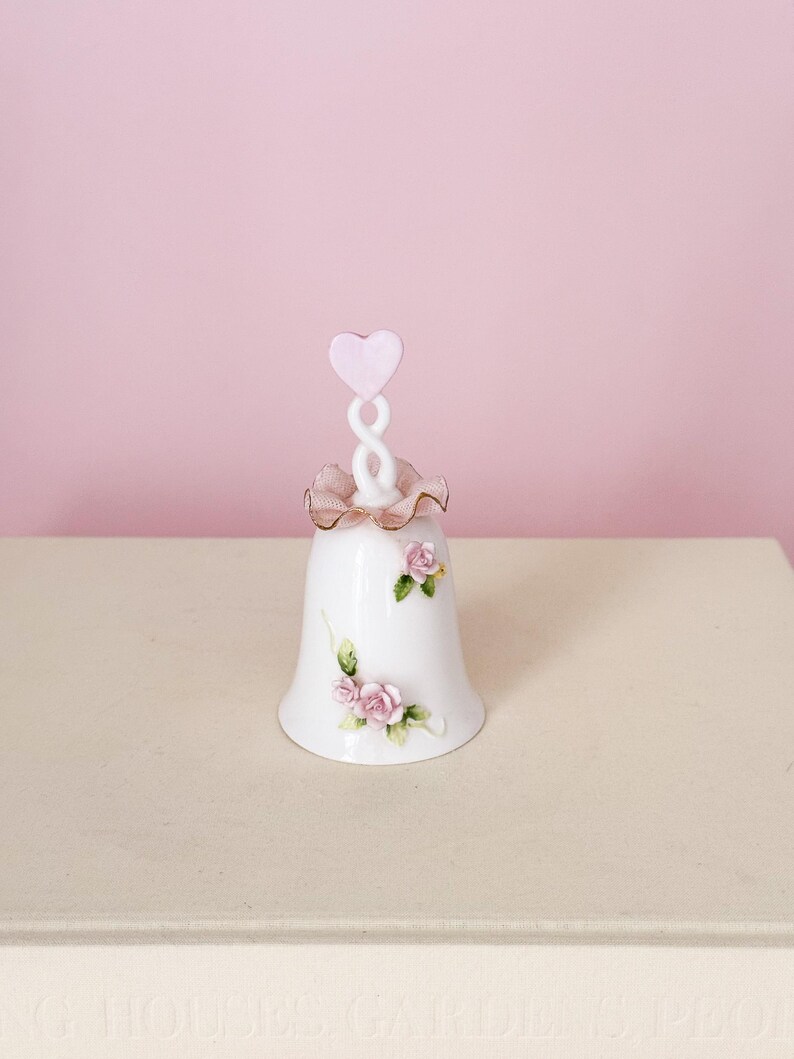 Puede incluir: Campana de porcelana blanca con detalles florales de rosas rosas. La campana tiene un asa en forma de coraz&oacute;n rosa y un ribete con volantes. Un art&iacute;culo decorativo, probablemente para exhibici&oacute;n o colecci&oacute;n.