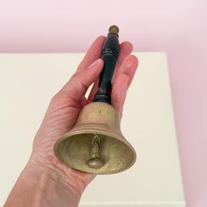 Puede incluir: Una campana de lat&oacute;n con un mango de madera negro y una peque&ntilde;a bola de lat&oacute;n en la parte superior. La campana se sostiene en una mano contra una superficie blanca y un fondo rosa. La campana es de dise&ntilde;o cl&aacute;sico.