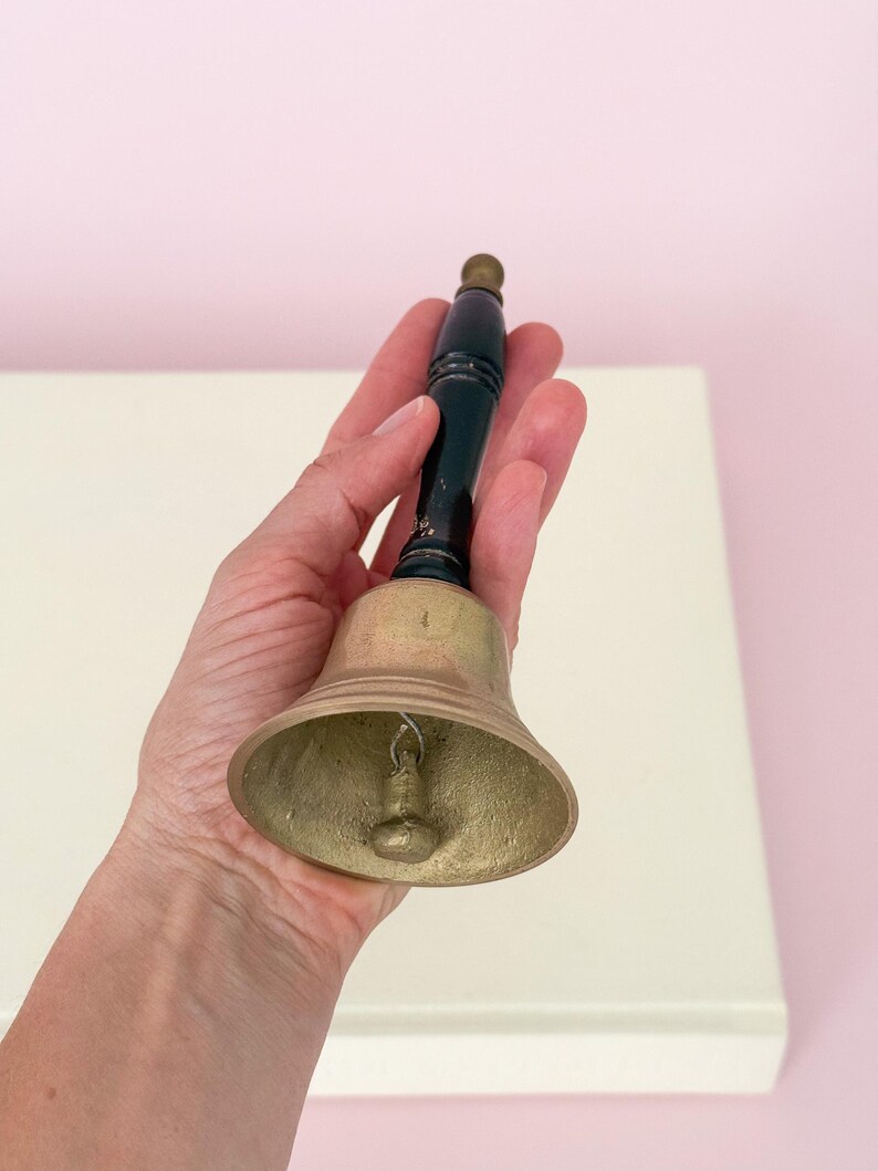 Puede incluir: Una campana de lat&oacute;n con un mango de madera negro y una peque&ntilde;a bola de lat&oacute;n en la parte superior. La campana se sostiene en una mano contra una superficie blanca y un fondo rosa. La campana es de dise&ntilde;o cl&aacute;sico.