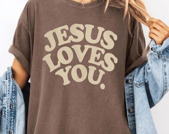 Puede incluir: Camiseta color espresso con el texto "JESUS LOVES YOU" en una fuente retro color crema. La camiseta tiene mangas enrolladas y un ajuste relajado. Una chaqueta vaquera está sobre los hombros.