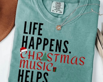 May include: A seafoam green t-shirt with the phrase "LIFE HAPPENS. CHRISTMAS MUSIC HELPS." in black and red lettering. A Santa hat, Christmas tree, and gift graphic are included. A silver necklace with a pendant is draped across the shirt.
