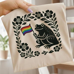 May include: A beige tote bag featuring a black illustration of a frog holding a rainbow flag. The frog is surrounded by a square floral border. The bag is held by a person.