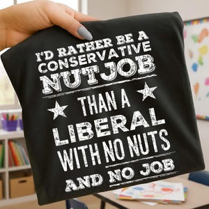 May include: Black t-shirt with white text. The text reads "I'D RATHER BE A CONSERVATIVE NUTJOB THAN A LIBERAL WITH NO NUTS AND NO JOB". The shirt is held up by a person.