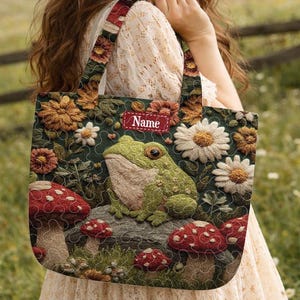 May include: A floral tote bag featuring a frog, mushrooms, and daisies. The bag has a dark green background with a quilted design. The handles and a small tag with the word "Name" are visible.