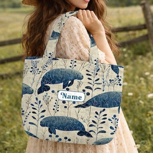 May include: A beige tote bag featuring a pattern of blue manatees and botanical elements. The bag has a rectangular shape with two handles and a label that says "Name". The bag is carried over the shoulder.