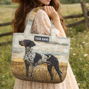 May include: A tote bag featuring a German Shorthaired Pointer dog design. The bag has a light blue and beige background with the text "YOUR NAME" on a black label. The dog is black and white, standing in a field.