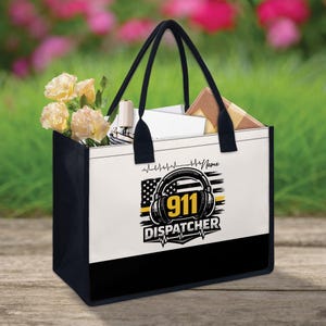 May include: A black and white tote bag with black handles. The bag features a design with the text "911 DISPATCHER" and a graphic of headphones over an American flag. The bag is filled with yellow flowers and other items.