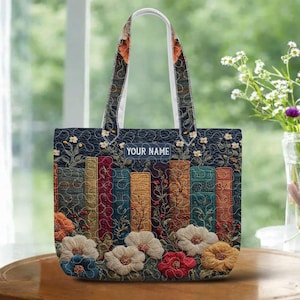 May include: A navy blue tote bag with a floral and book design. The bag features a row of colorful, textured books and embroidered flowers in shades of white, red, and yellow. The text "YOUR NAME" is displayed on the front.
