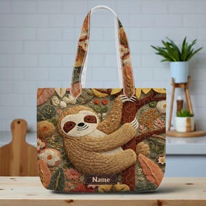 May include: Embroidered tote bag featuring a detailed sloth design hanging from a tree branch. The bag has a cream-colored handle and a floral pattern. The word "Name" is displayed on the bottom of the bag.