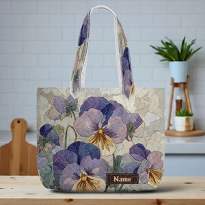 May include: A tote bag featuring a floral design of purple and yellow pansies. The bag has a light-colored background and matching handles. A small brown tag with the word "Name" is visible on the front of the bag.