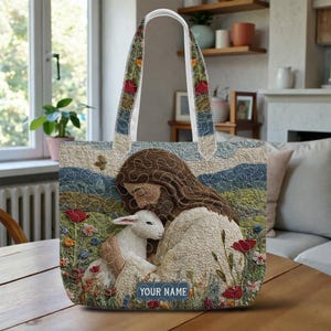 May include: A quilted tote bag featuring a serene scene of a figure embracing a lamb amidst a field of wildflowers. The bag has a floral patterned strap and the text "YOUR NAME" on the bottom. The color palette includes blues, greens, and reds.