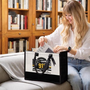 May include: A black and white tote bag with a graphic of a headset, American flag, and the text "911 DISPATCHER". The bag has black handles and a black bottom. The bag is being filled with a book.