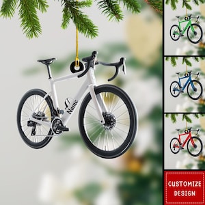 May include: A white bicycle ornament with black wheels and handlebars hangs from a gold string. The word "Name" is printed on the frame. Additional ornaments in green, blue, and red are also shown. The image includes a "Customize Design" button.