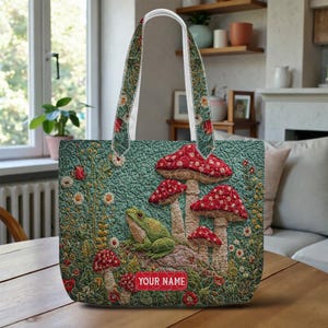 May include: A teal tote bag featuring a whimsical design of red-capped mushrooms, a green frog, and wildflowers. The bag has a quilted texture and floral patterned handles. The text "YOUR NAME" is displayed on a red rectangle.