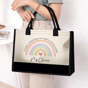 May include: A beige tote bag with black handles and a black bottom panel. The bag features a pastel rainbow design with the text "It takes a kind heart to inspire young minds" and the name "Ms. Olivia".