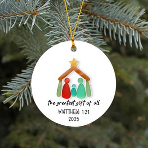 May include: White Christmas ornament with a gold string, featuring a colorful illustration of a family under a roof with a star. Text reads "the greatest gift of all" and "MATTHEW 1:21 2025".