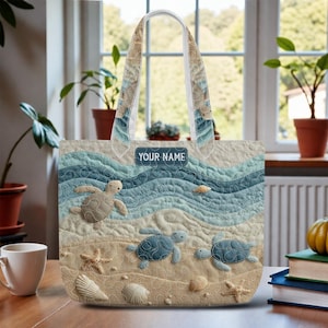 May include: A quilted tote bag featuring a beach scene with turtles, seashells, and starfish. The design includes blue waves and sand, with the text "YOUR NAME" on a label. The bag has a white handle and is set against a window background.
