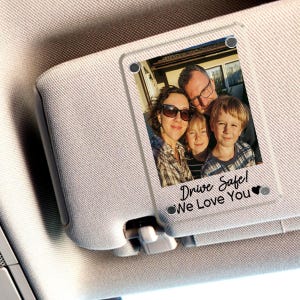 May include: A clear acrylic car visor photo charm with a family photo. The photo charm is attached to a beige car visor. The text "Drive Safe! We Love You" is printed on the charm.