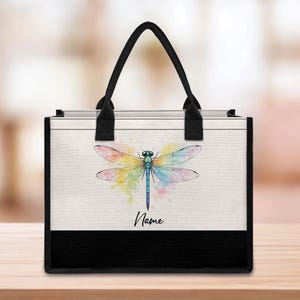 May include: A tote bag with a watercolor dragonfly design. The dragonfly has colorful wings and a blue body. The bag has a black base and handles, with the word "Name" printed below the dragonfly.
