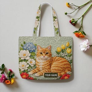 May include: A light green tote bag featuring a detailed illustration of an orange tabby cat lying among flowers. The bag has white floral patterned handles and the text "YOUR NAME" on the bottom.